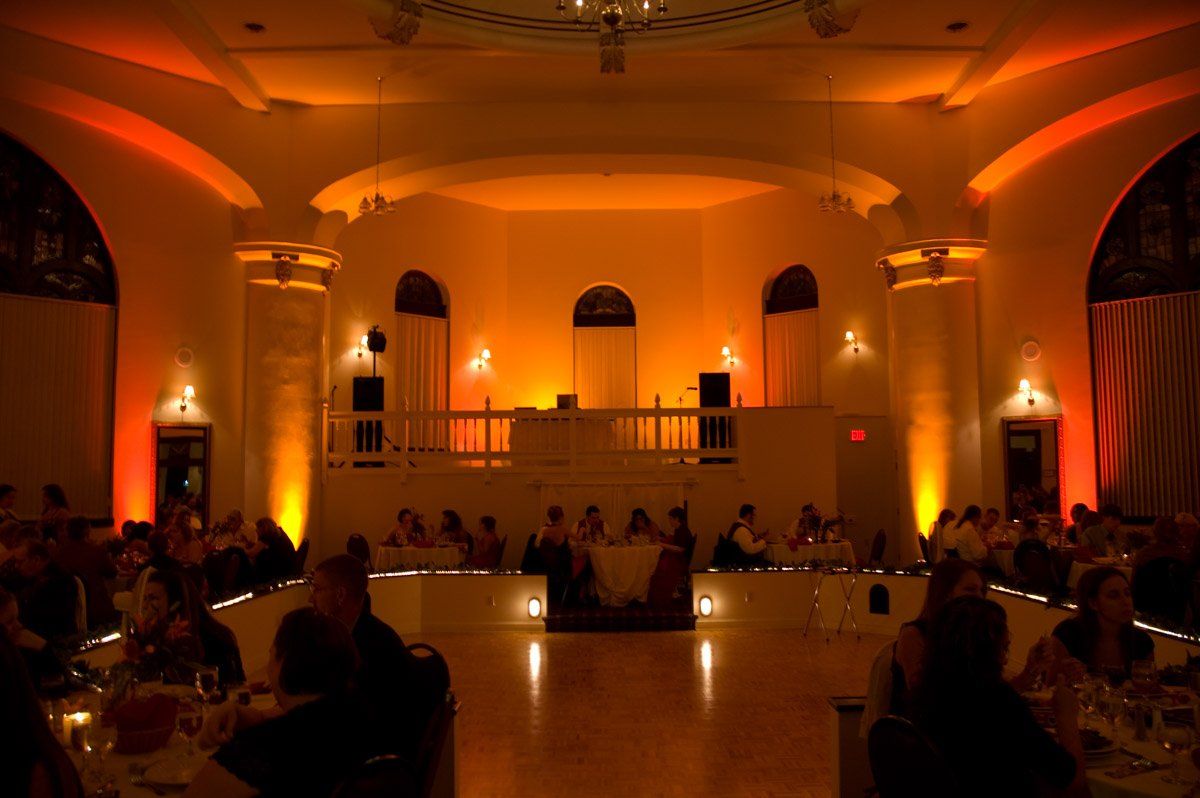Ballroom with amber uplighting, people seated at tables. Stage with a DJ setup.
