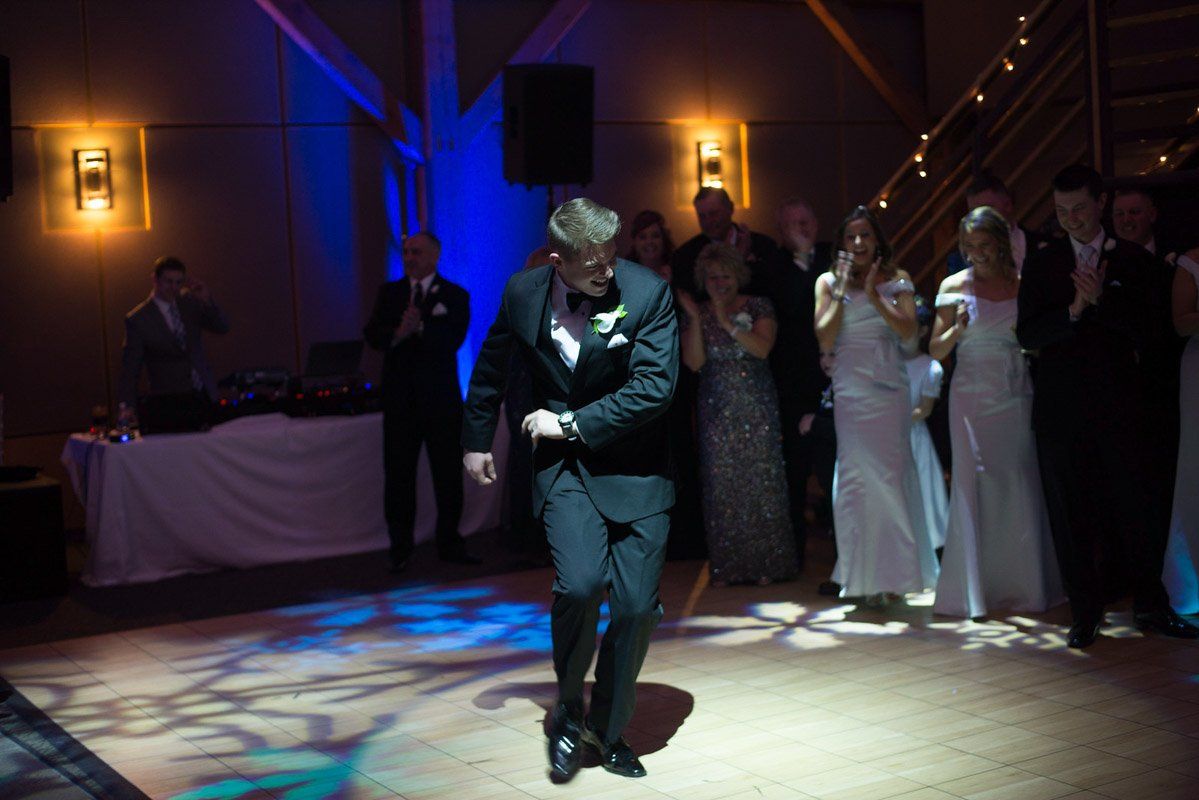 A young man in a tuxedo dances on a dance floor, surrounded by people in formal attire, clapping.