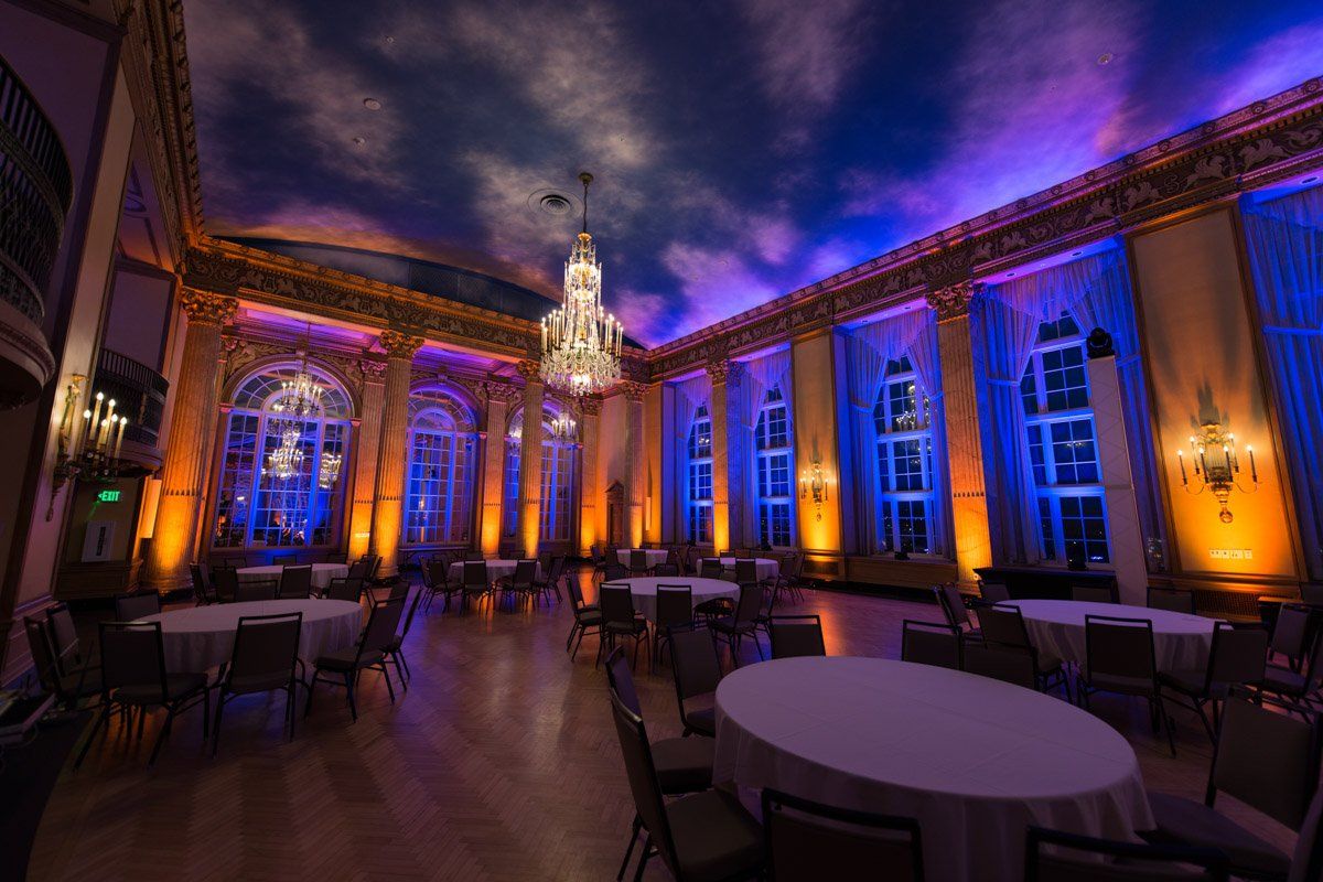 Ornate ballroom lit with orange and blue lights, tables set for a gathering, dramatic painted ceiling.