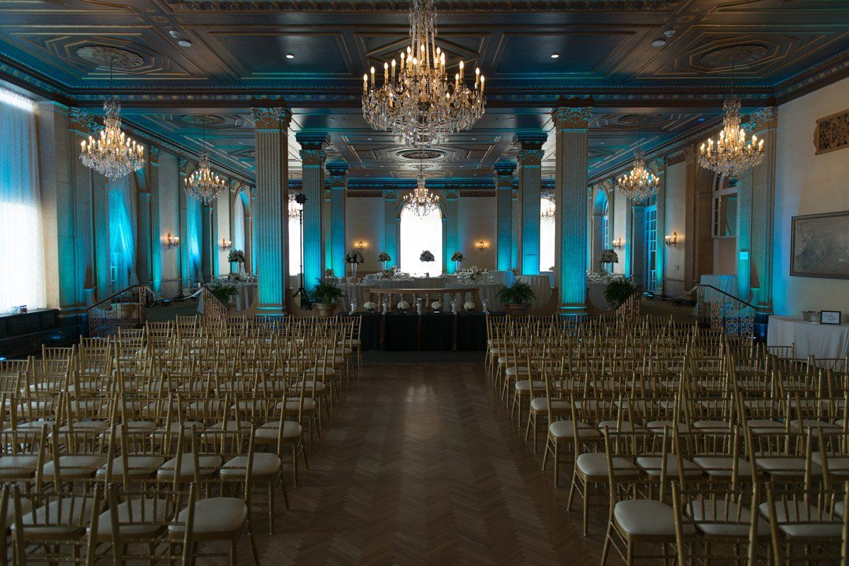 Formal room set for an event with gold chairs, ornate columns, chandeliers, and blue lighting.