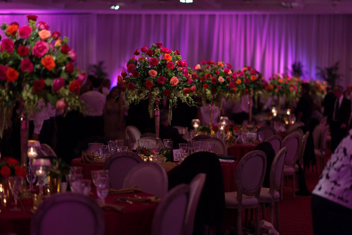 Elegant banquet hall with round tables, tall flower arrangements, and purple lighting.