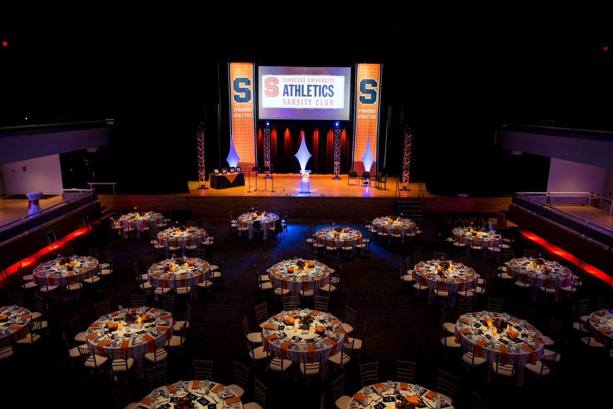 Banquet hall with round tables, stage with banners, orange and blue lighting.