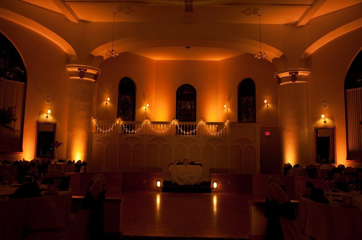 An ornate room with warm orange lighting, set up for an event with a stage, decorated ceiling, and tables.