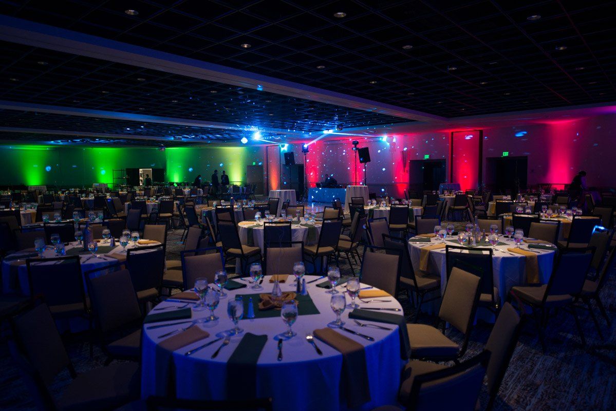 Banquet hall with round tables, decorated with green and white, and colorful lighting.