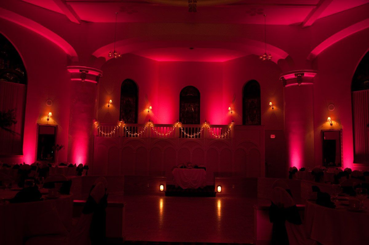 Pink-lit ballroom; stage with decorative balcony, three arched windows. Tables and chairs in shadows.