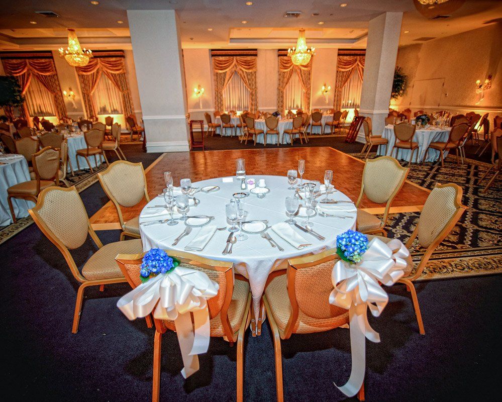 Round table set for a wedding reception with chairs adorned with bows; chandeliers hang overhead.