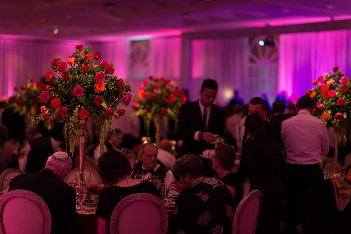 Elegant dinner party with tall red floral arrangements, pink lighting, and people seated at tables.