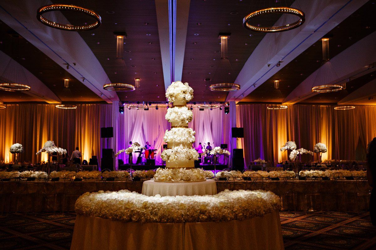 Elegant wedding reception: tiered cake center stage, surrounded by floral arrangements, tables, band, warm lighting.