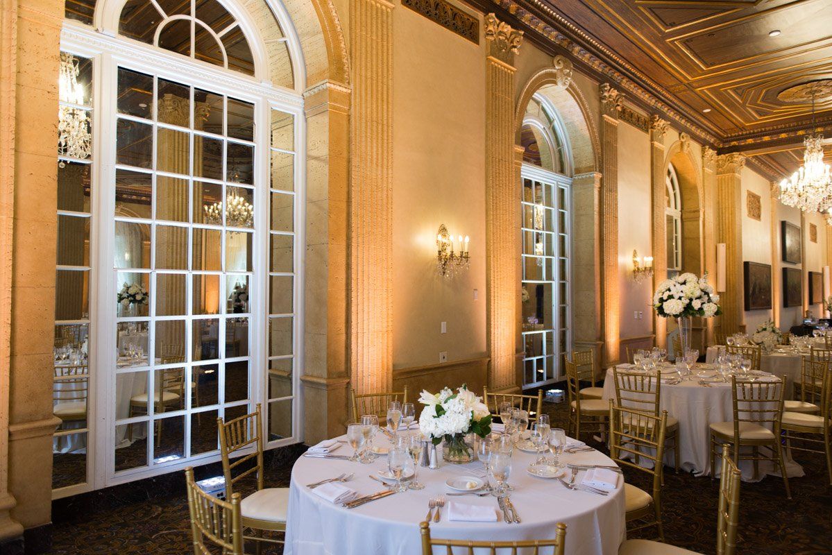 Elegant banquet hall with round tables set for a formal event. White flowers, gold chairs, and large windows.