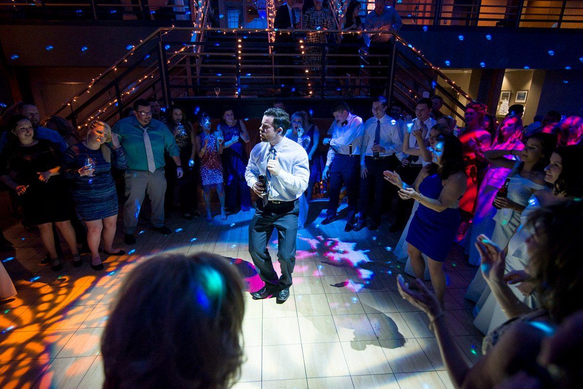 Man dances on a lit dance floor surrounded by people at a party, under colorful disco lights.