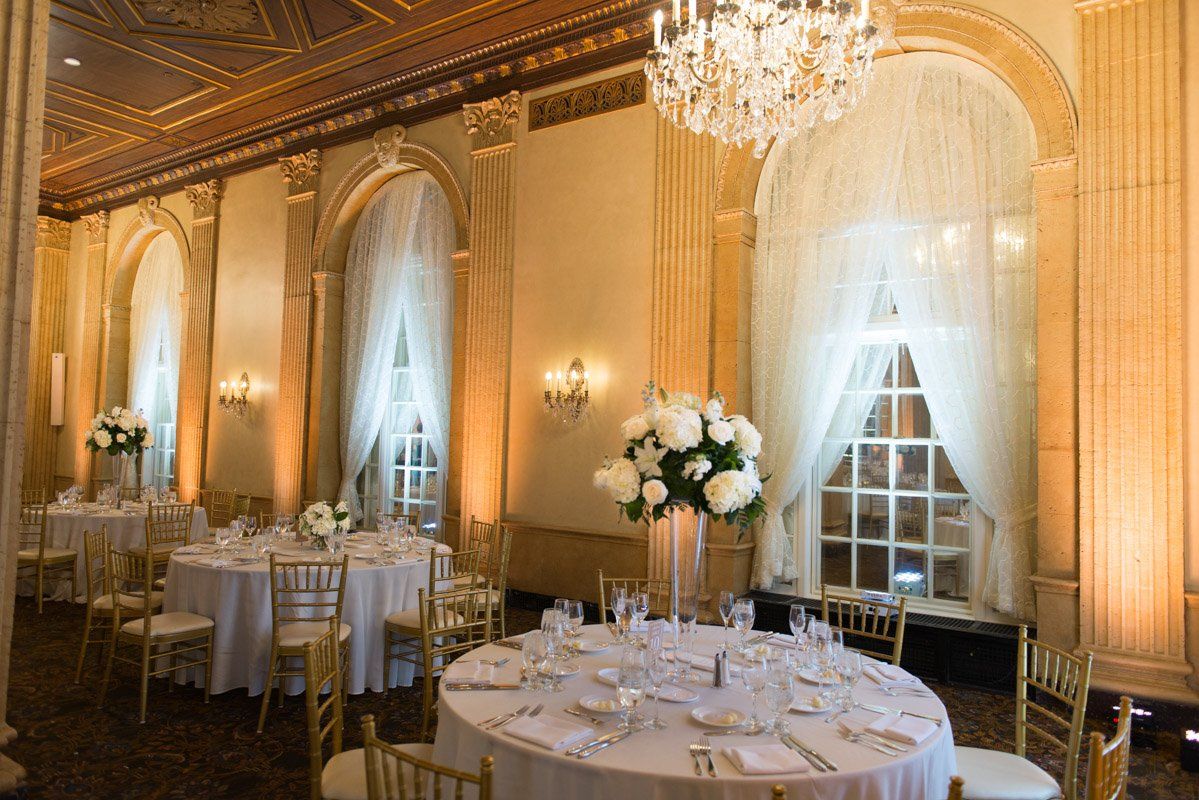 Elegant ballroom with round tables set for a formal event; featuring white floral centerpieces, gold chairs, and a large chandelier.