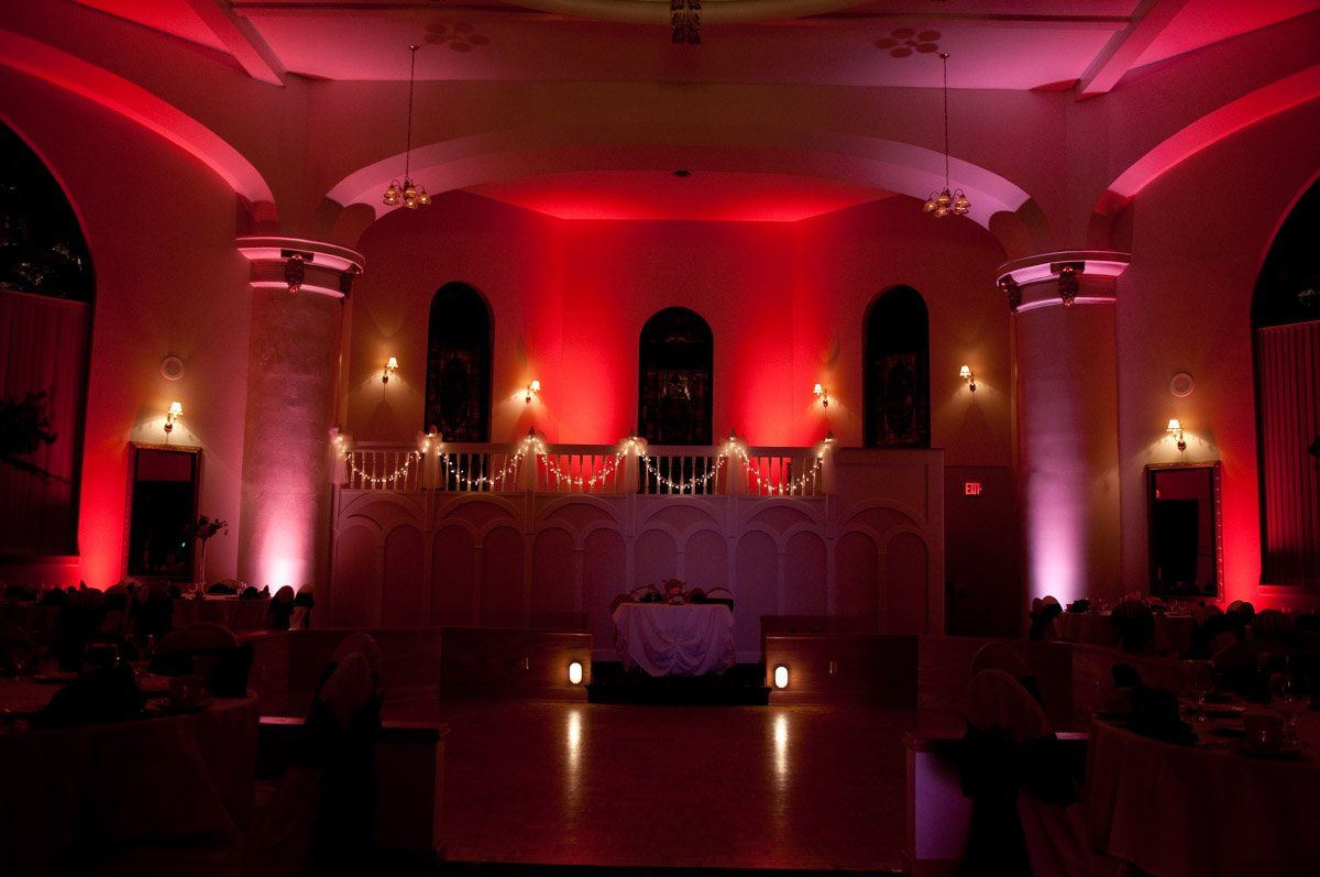 Red uplighting illuminates a wedding reception hall; stage with floral arrangement.