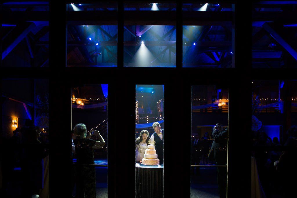 Wedding cake cutting through glass windows; couple, guests, and blue-lit party scene.