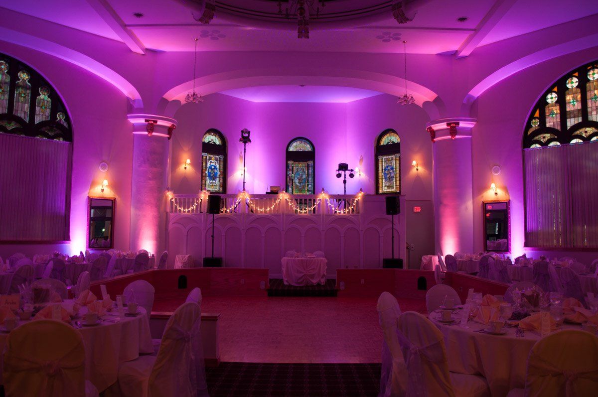 A wedding reception hall bathed in pink light, tables set, stage with white decor and stained glass windows.