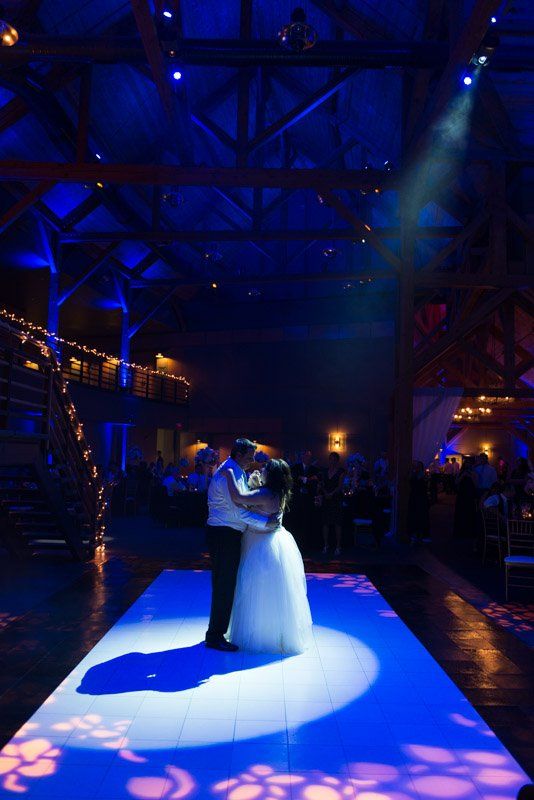 Couple dances in a spotlight on a blue-lit dance floor at a wedding reception. Dark room, guests in background.