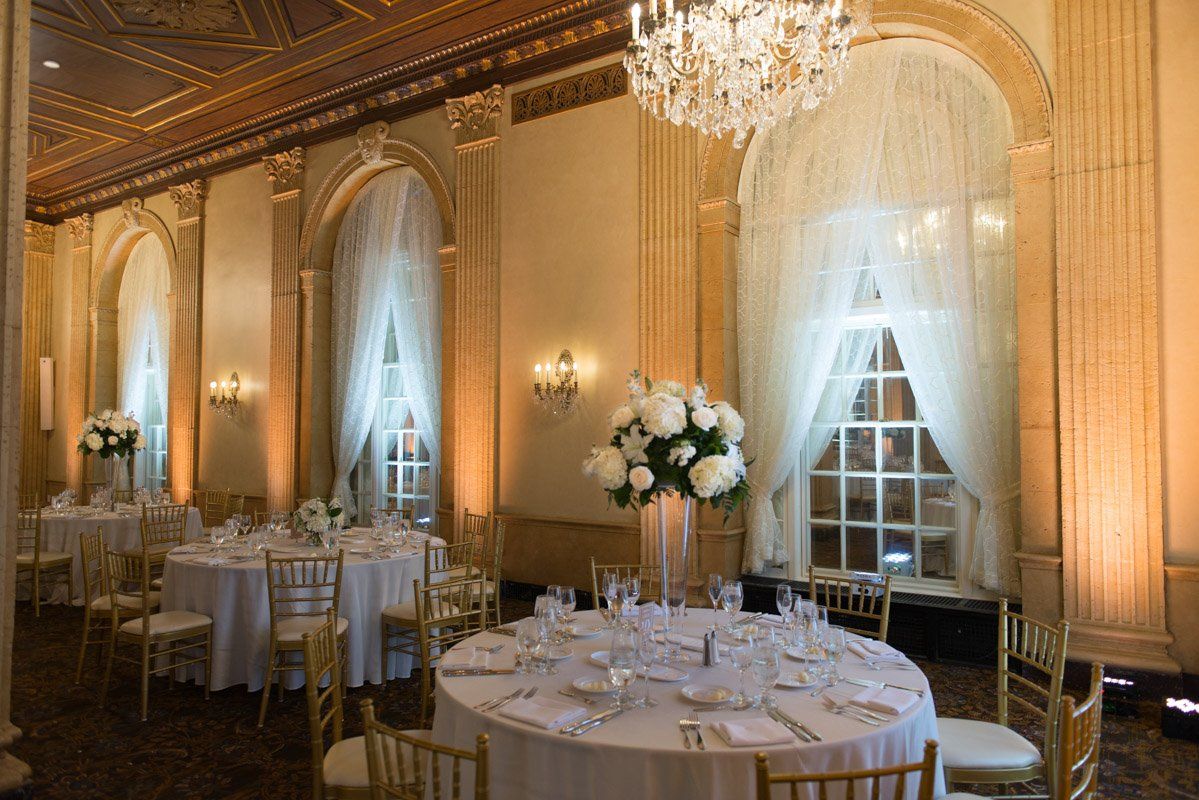 Elegant banquet hall with round tables set for a wedding. White flowers, gold chairs, and soft lighting.
