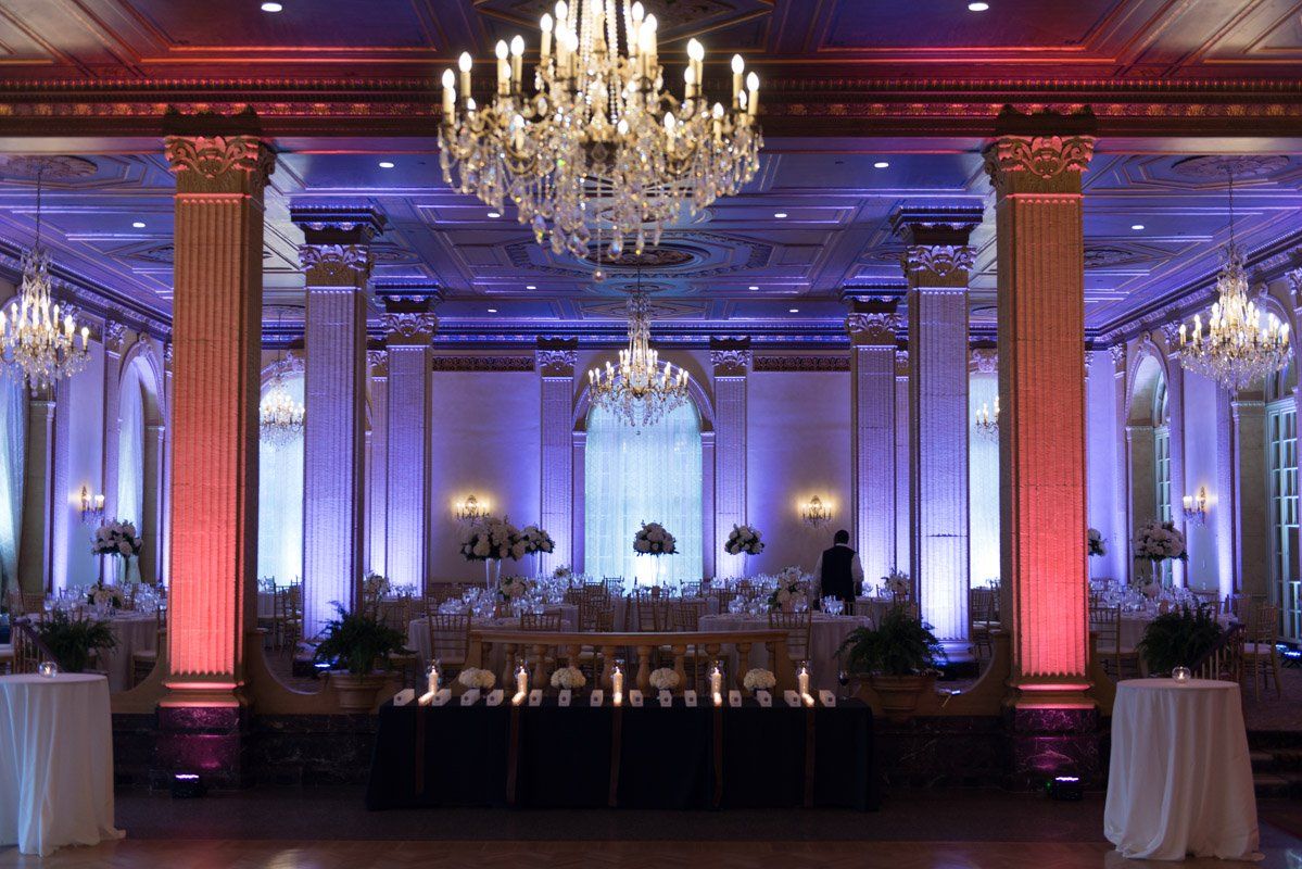 Elegant ballroom with crystal chandeliers, columns, and tables set for an event. Purple and red lighting.