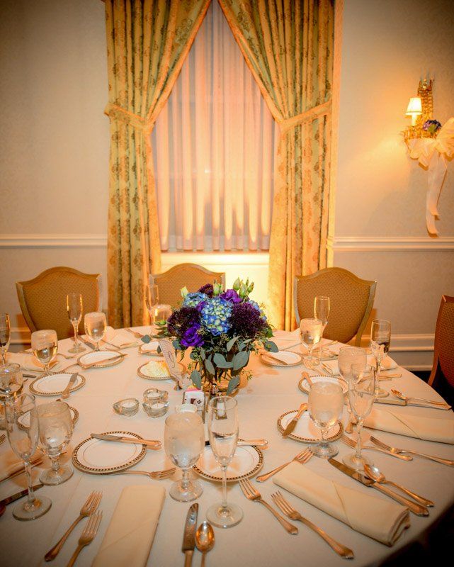 Elegant dining table set for guests, floral centerpiece, window with gold curtains, light tan chairs.