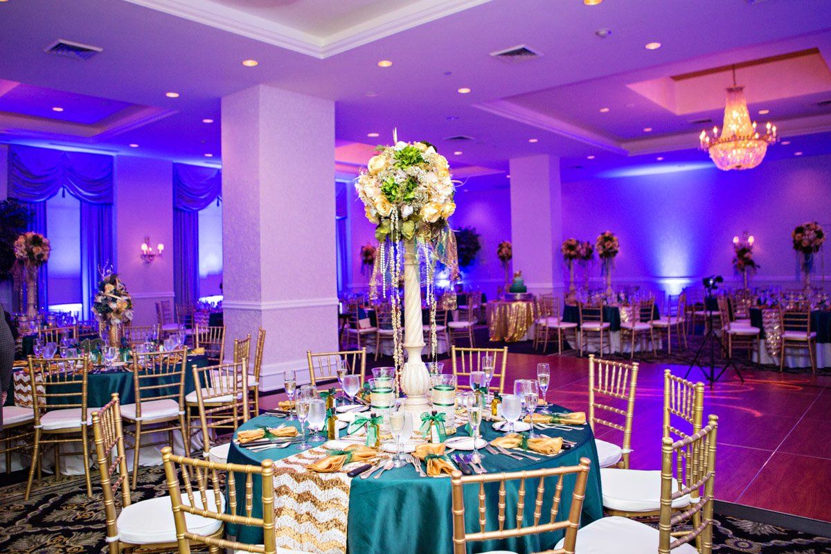 Elegant ballroom with round tables, teal and gold decor, and floral centerpieces, lit with purple and gold hues.