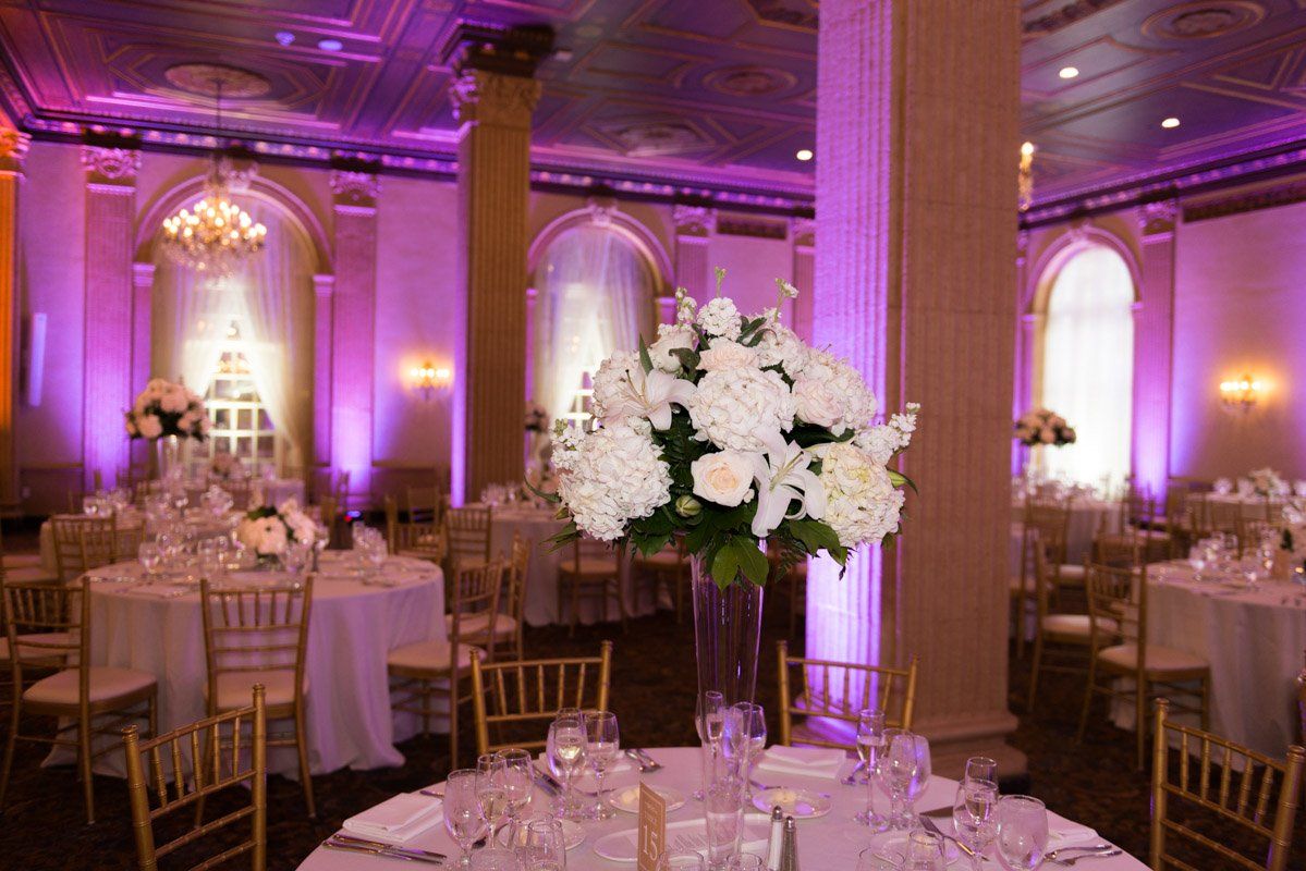 Wedding reception hall with round tables, gold chairs, floral centerpieces, and purple and orange lighting.