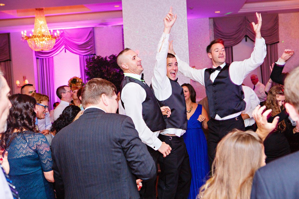 Wedding reception dance floor, three men in vests celebrating with arms raised.