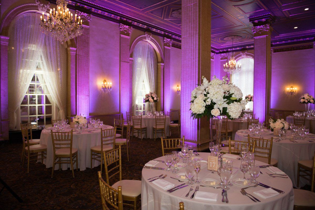 Elegant banquet hall with round tables, white tablecloths, floral centerpieces, and purple uplighting.