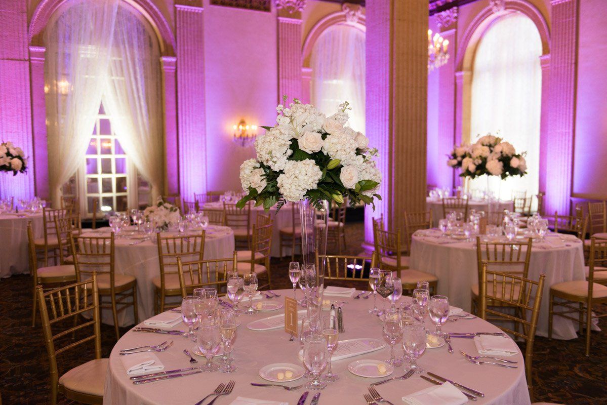 Elegant ballroom decorated for a wedding with round tables, floral centerpieces, and purple lighting.