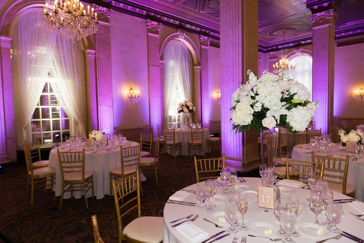 Elegant ballroom decorated for an event. Round tables with white linens, floral centerpieces, and purple lighting.
