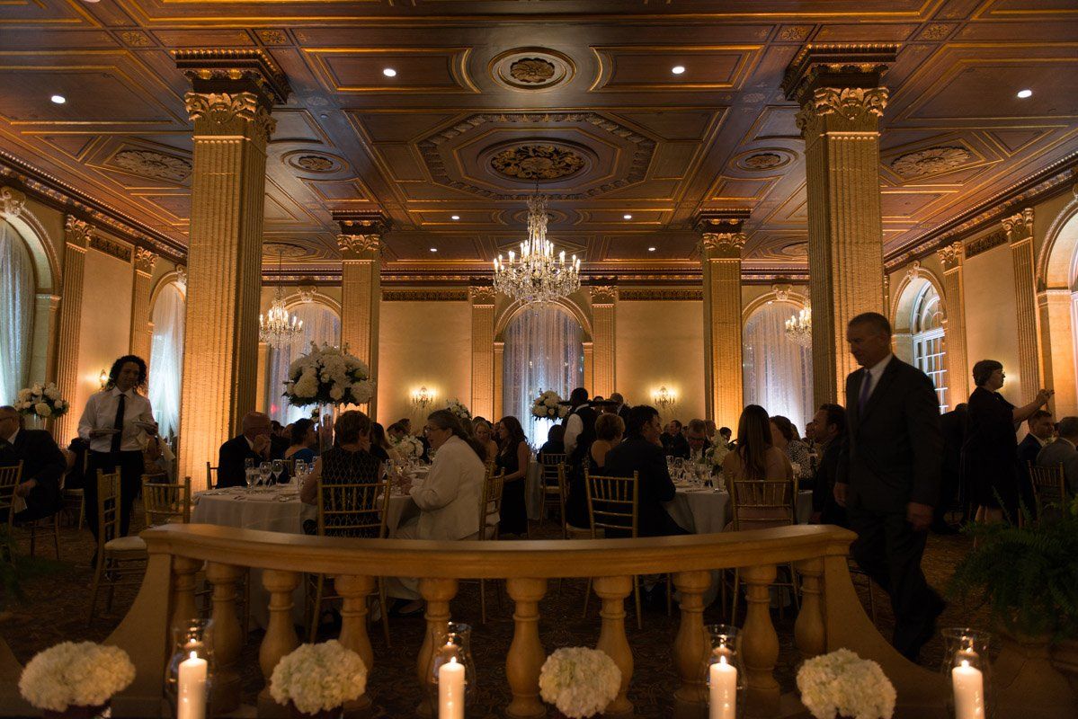 Elegant ballroom with chandelier, tables, guests, and a man walking; warm lighting.