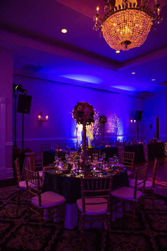 Formal dining room with round tables, centerpieces, and gold chairs, lit with blue and purple lighting.