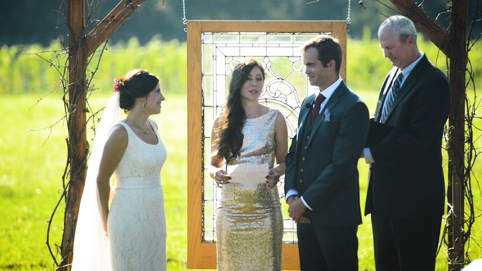 Wedding ceremony outdoors: Bride, groom, bridesmaid, officiant stand before a stained glass backdrop, sunny day.