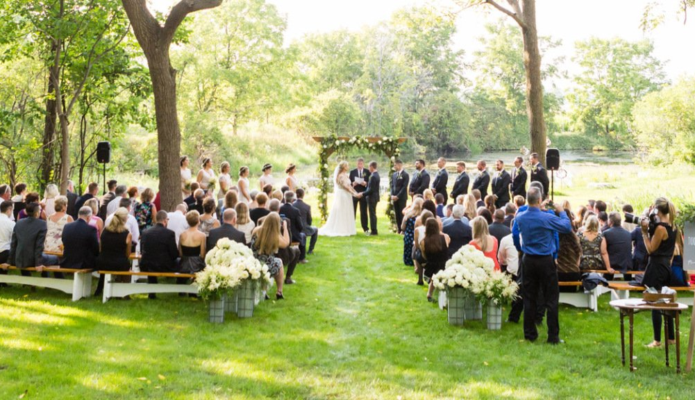 Outdoor wedding ceremony: Bride and groom at altar, guests seated on benches, green grass and trees.