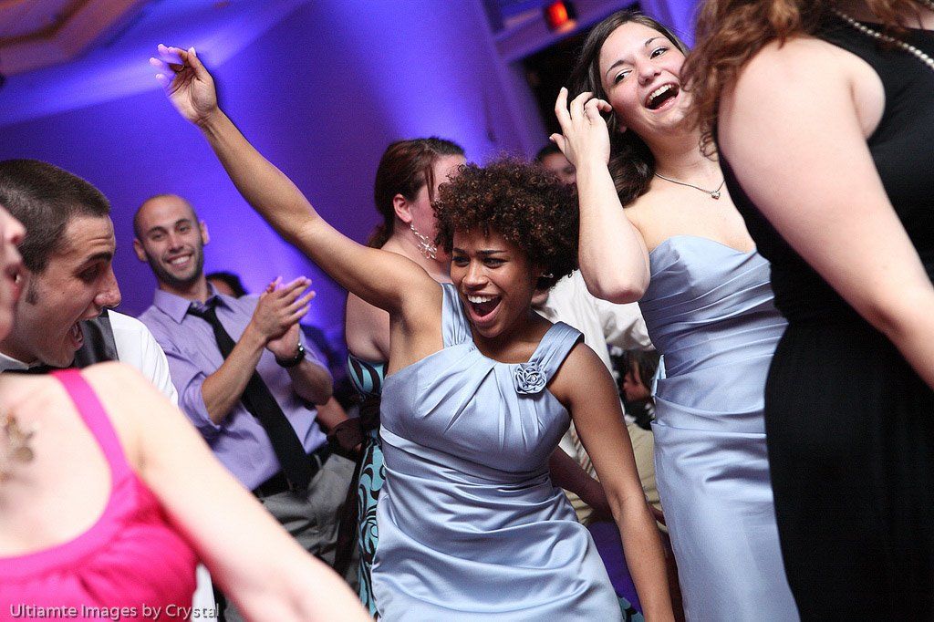 People dancing at a party. A woman in a blue dress raises her arms, others smile and clap under blue lights.