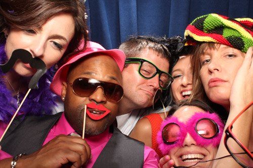 Group of friends in a photo booth, smiling and posing with props like mustaches and glasses.