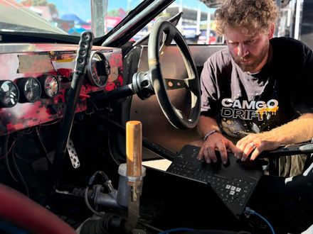 Cameron Ombler-Vrey wearing a CamGoDrift shirt is working on a laptop in a car