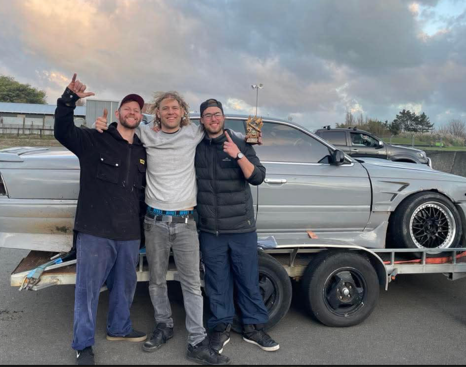 Three men are posing for a picture in front of a car on a trailer.