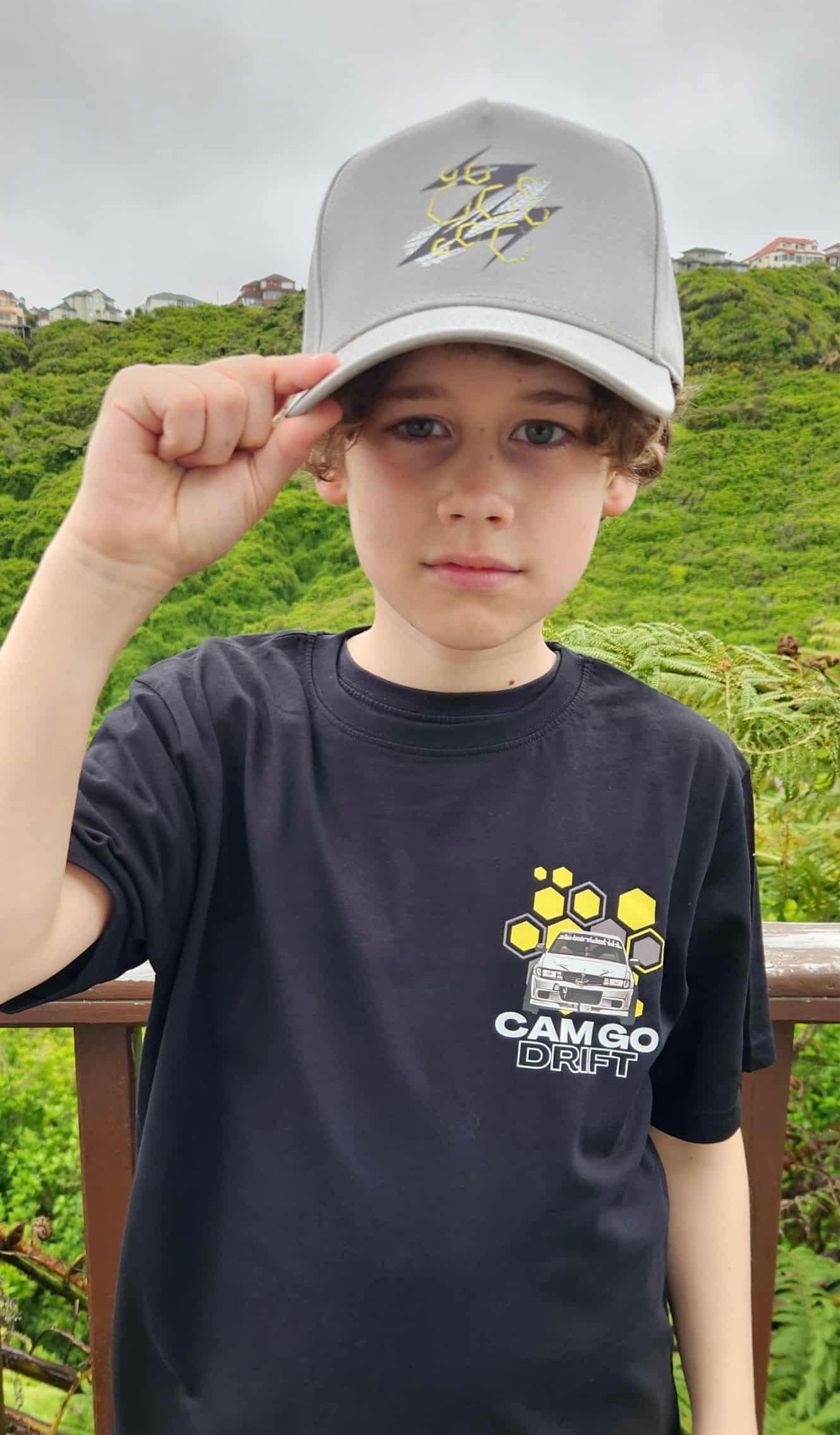 Boy in gray hat and black shirt with graphic, touching cap, outdoors.