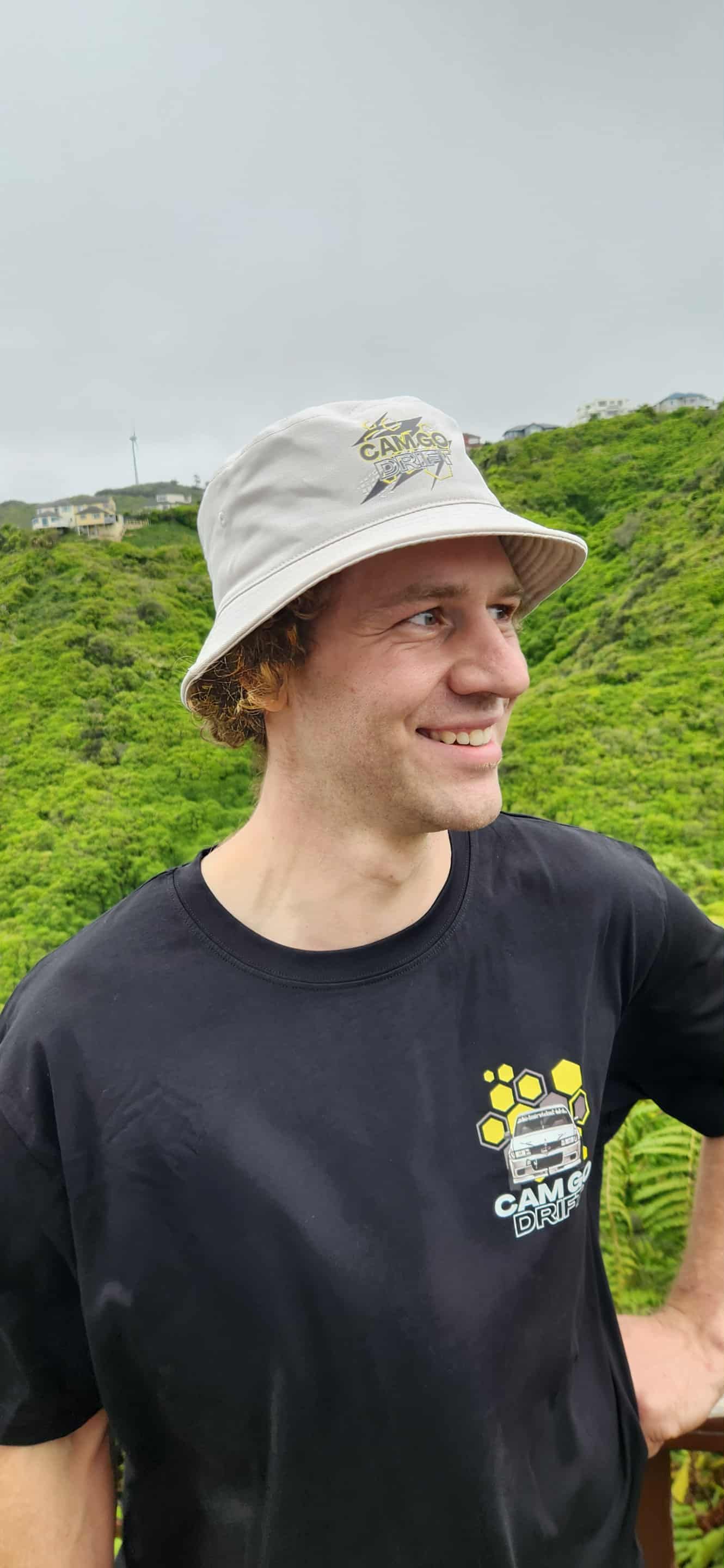 Man in a black t-shirt and bucket hat smiles, standing outdoors with greenery in the background.