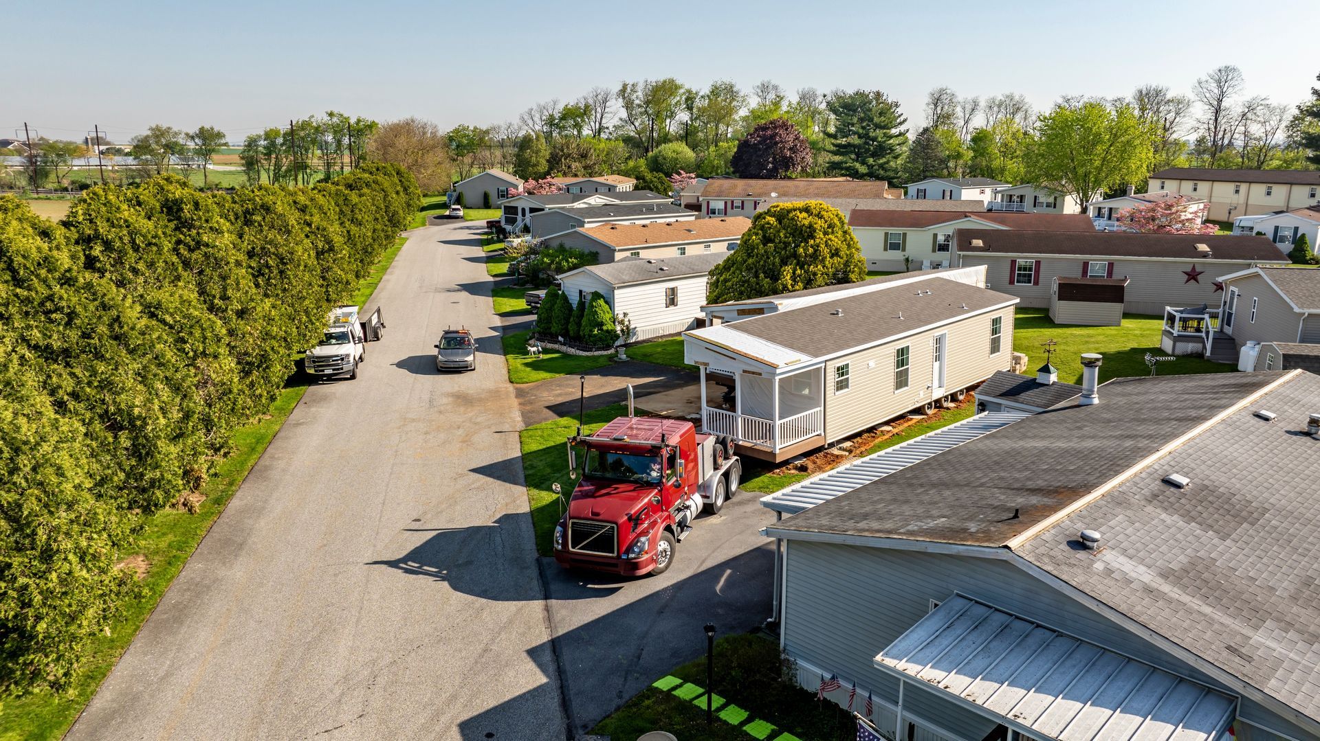 A red semi-truck moves a manufactured home through a residential street in a mobile home park on a sunny day.