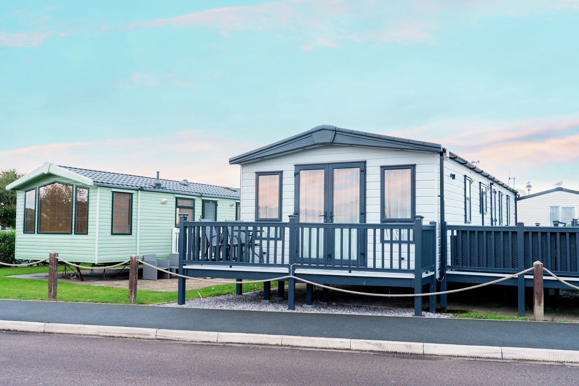 Two light-colored mobile homes with decks sit side-by-side on a grassy lot along a paved road under a cloudy sky.