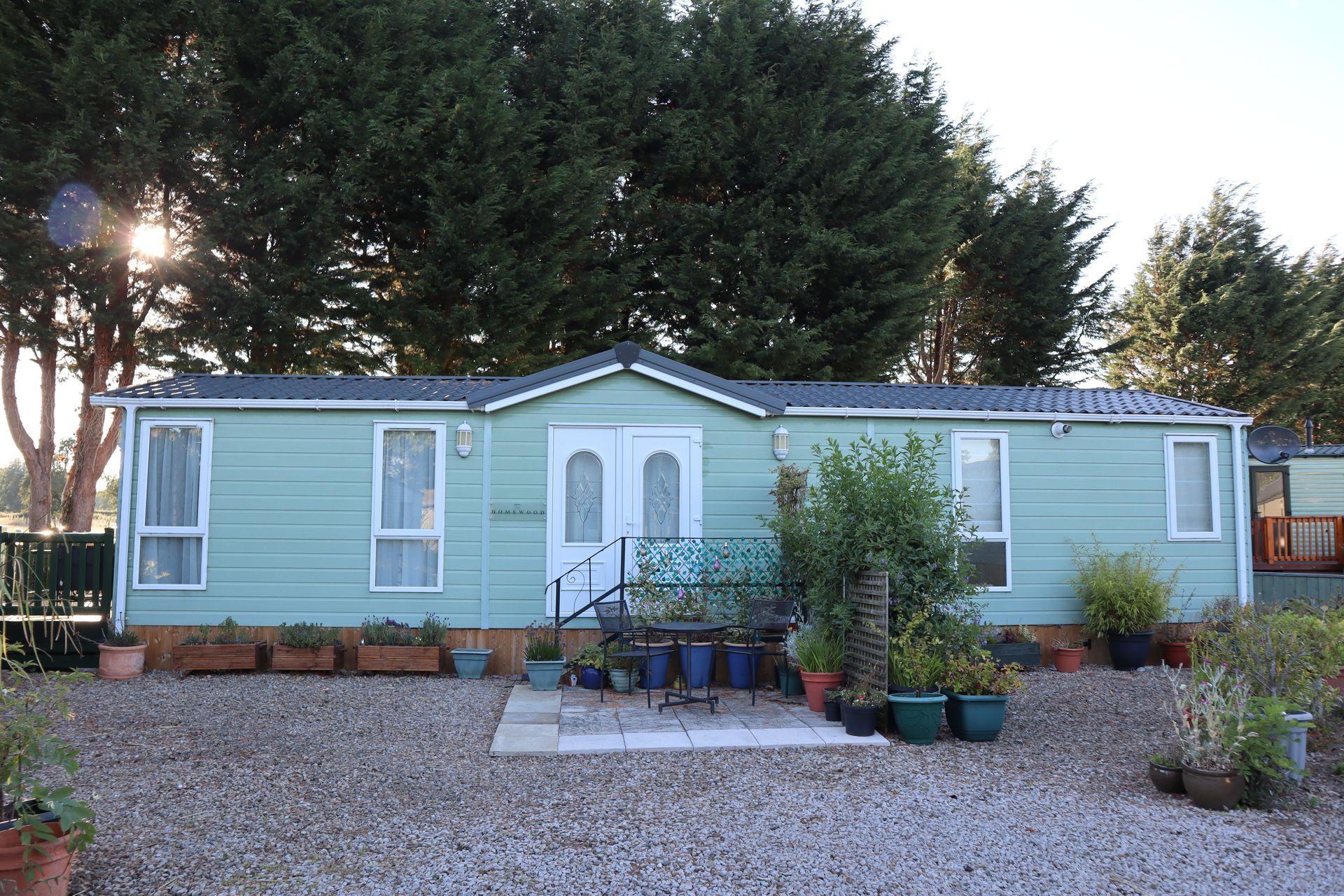 A light green mobile home with white trim and French doors, situated in a gravel yard with potted plants and trees.