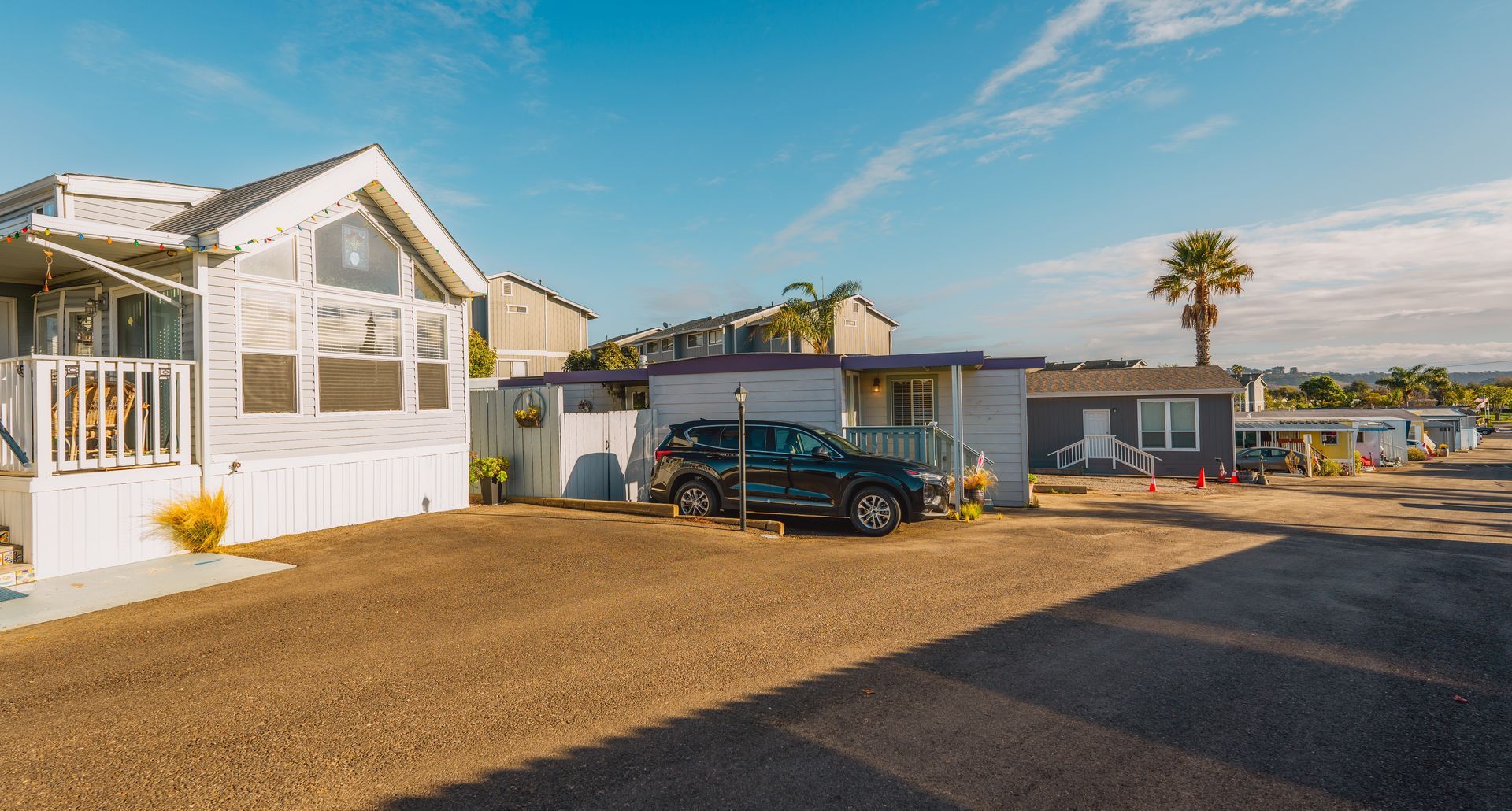 A dark SUV parked in front of light-colored mobile homes on a sunny, gravel-covered street.