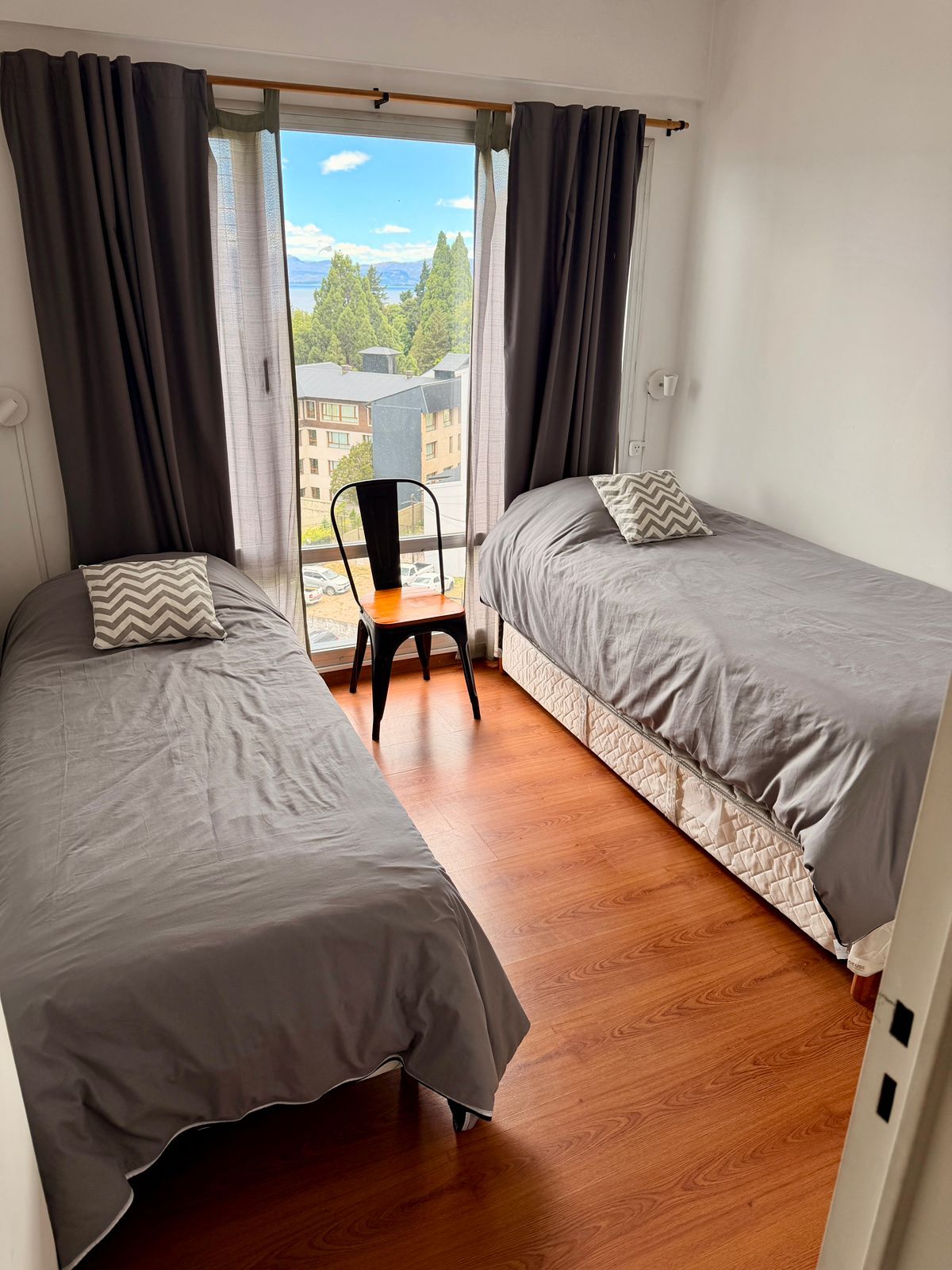 Dos camas individuales con ropa de cama gris, una silla y una ventana con vista.