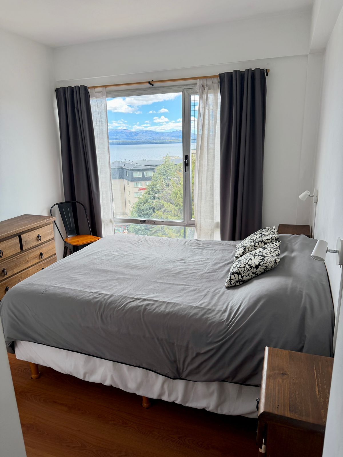 Dormitorio con cama, cómoda, silla y gran ventanal con vista a un cuerpo de agua y montañas.