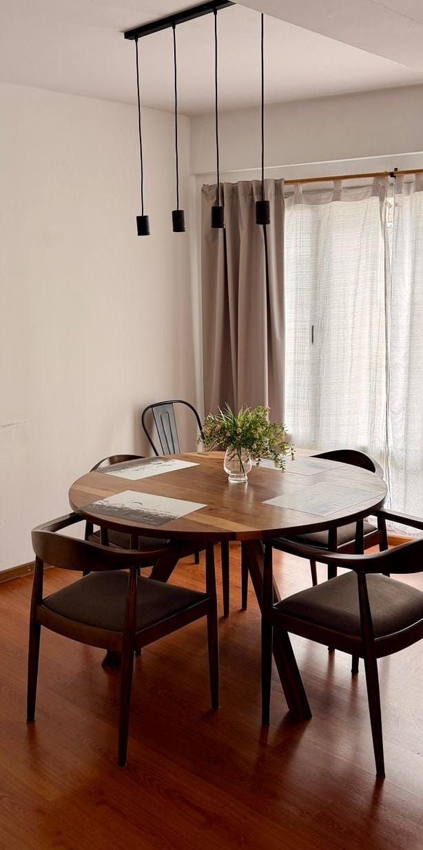 Comedor con mesa redonda de madera, cuatro sillas oscuras, una planta y lámparas colgantes negras.
