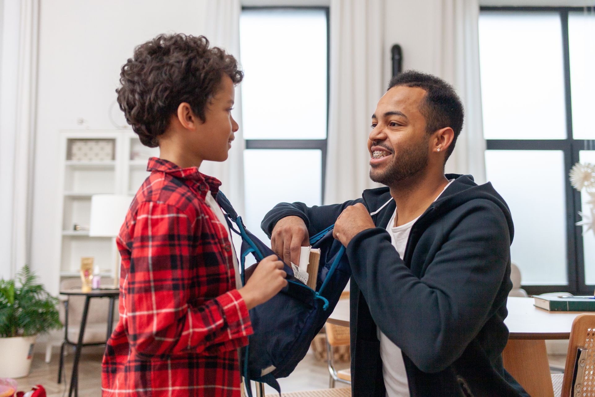 A father gets his son ready for school at home by putting a book in his backpack. A father gets his son ready for school at home by putting a book in his backpack.