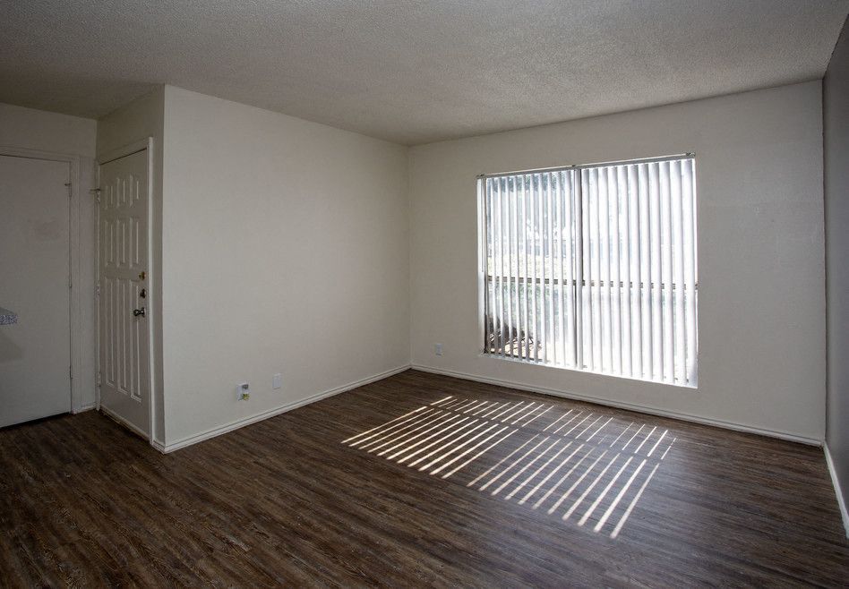 Empty living room with wood-look flooring and a large window with vertical blinds letting in sunlight.