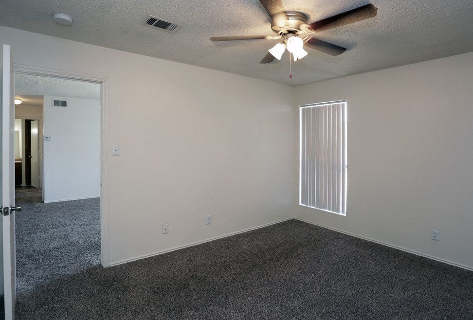 Empty apartment bedroom with ceiling fan, window with vertical blinds, and gray carpet.