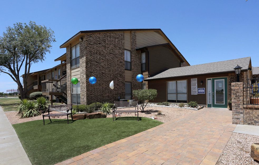 Exterior view of a brick apartment community with benches, landscaping, and a paved entry.