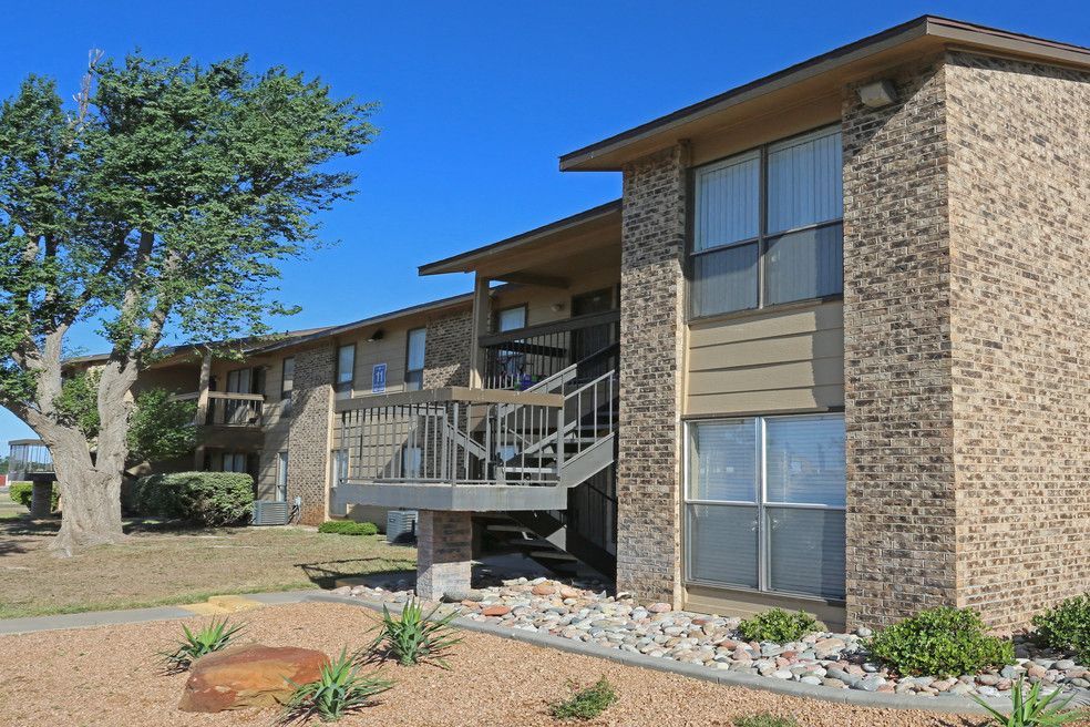 Exterior view of a brick apartment building with stairs, landscaping, and a large tree under a blue sky.